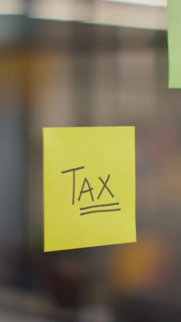 Vertical-Video-Close-Up-Of-Woman-Putting-Sticky-Note-With-Tax-Written-On-It-Onto-Transparent-Screen-In-Office